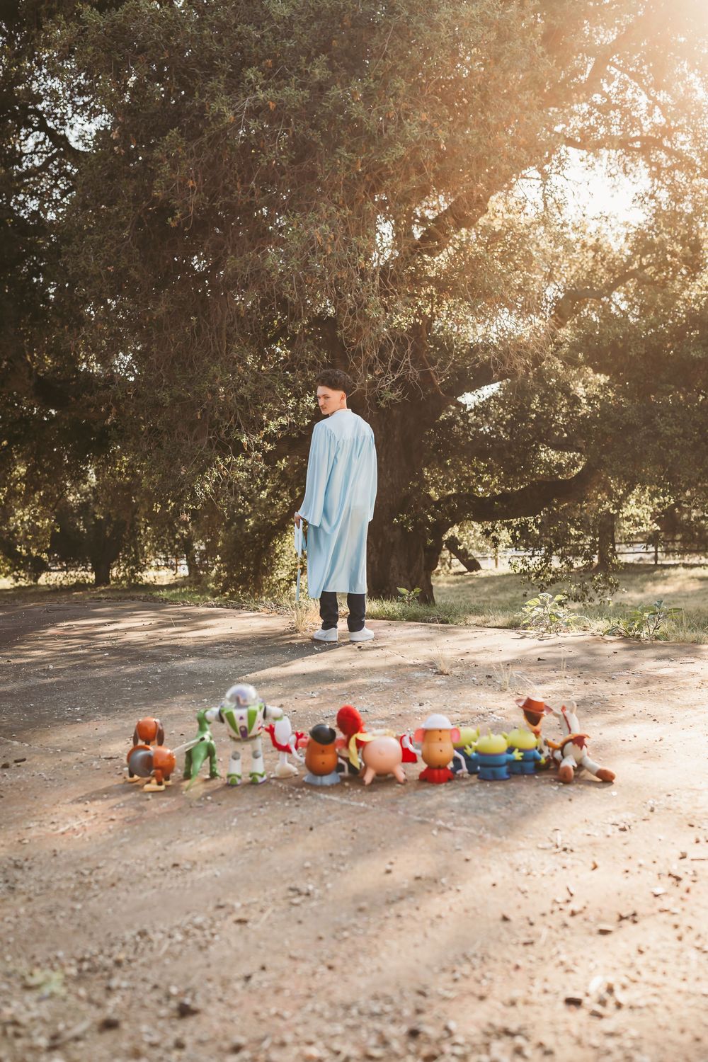 Graduate in blue gown stands on dirt path surrounded by Toy Story character figures in sunny park in yucaipa ca
