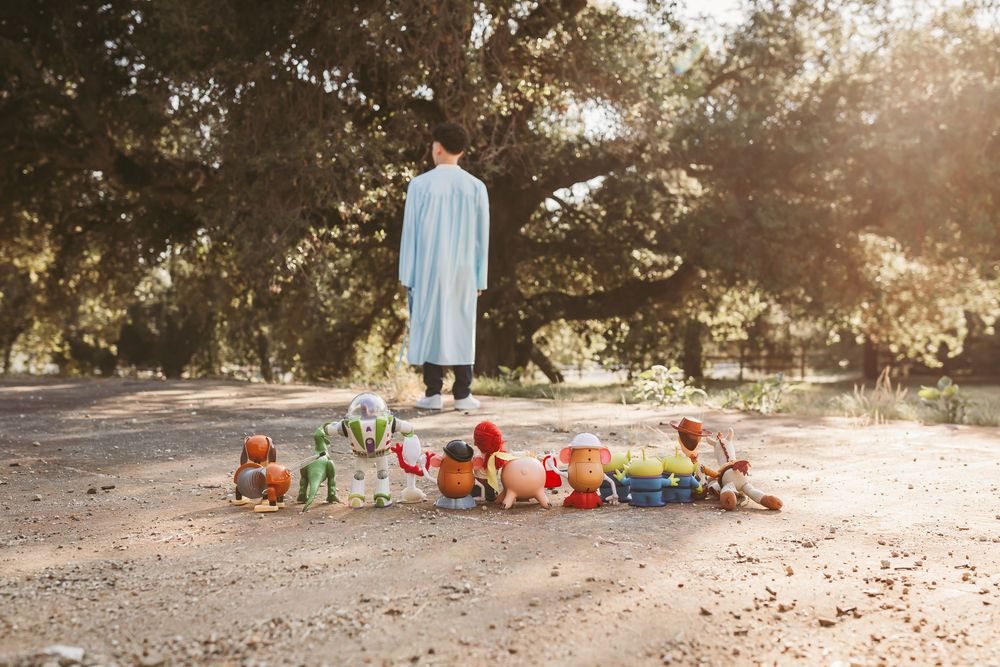 Child in blue gown stands among Toy Story character toys arranged on sandy ground beneath large oak trees for senior photos
