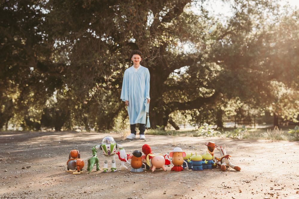 Person in blue robe stands among colorful Toy Story character plush toys on a dirt path under large trees.