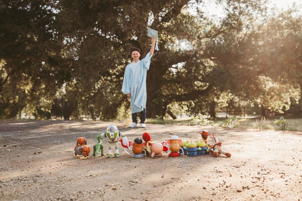 Graduate in blue gown jumps joyfully holding diploma above Toy Story toys on sunlit park path with heidi grace photography