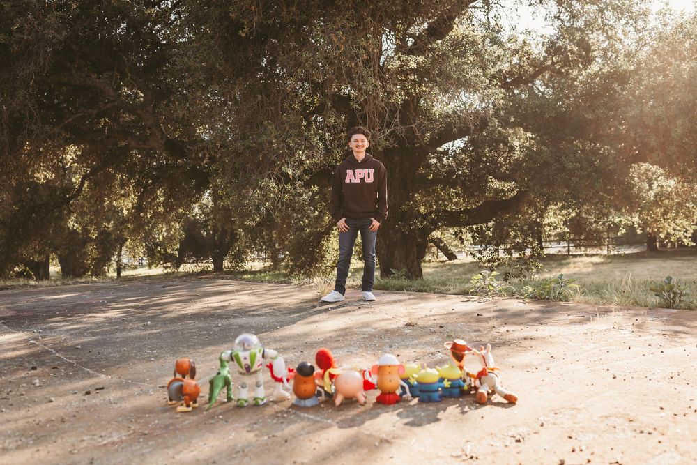 Person in APU hoodie standing on a sunlit path with colorful Toy Story character toys arranged in front.
