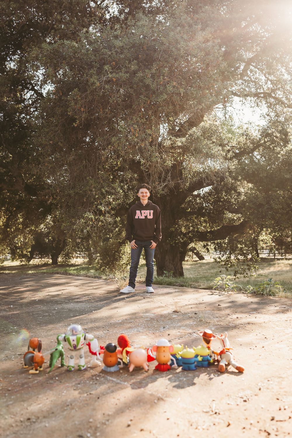 Person in APU hoodie standing outdoors on dirt path with Toy Story figurines lined up in front, large oak tree behind.
