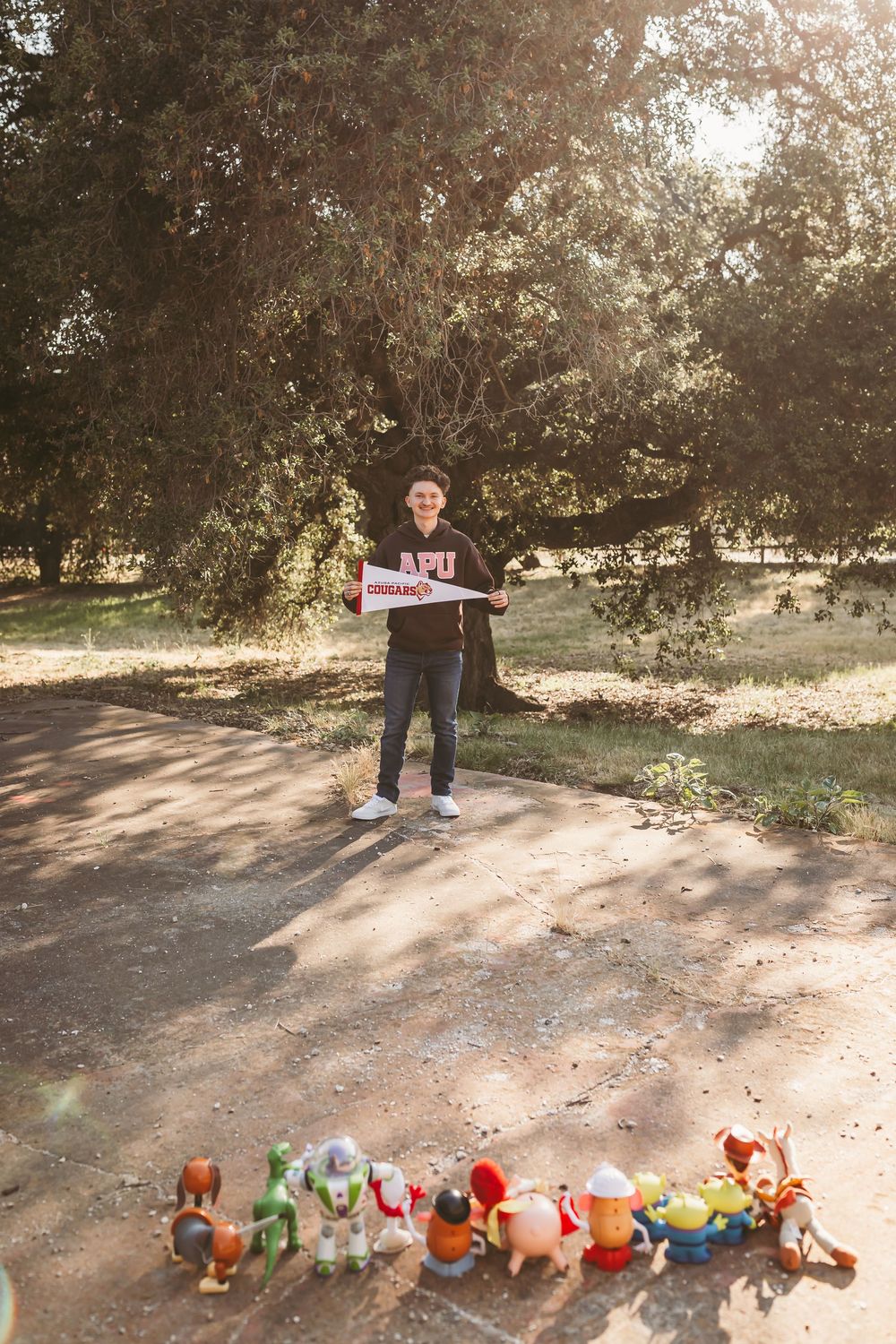 Teen holds an APU pennant flag while Toy Story figurines are lined up on a path in a sunny park.