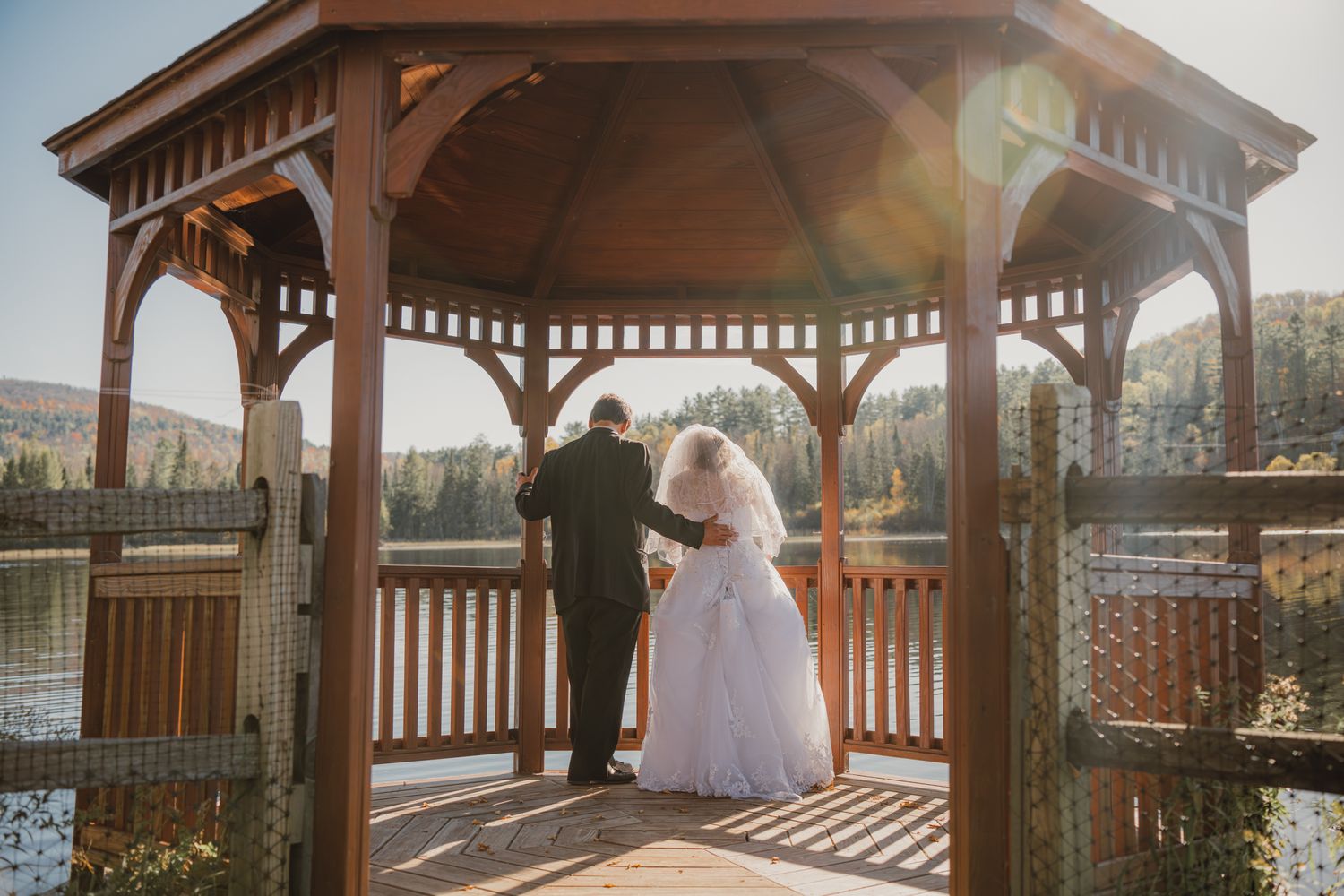 Bride and groom share a romantic moment in a wooden gazebo overlooking a serene autumn lake.