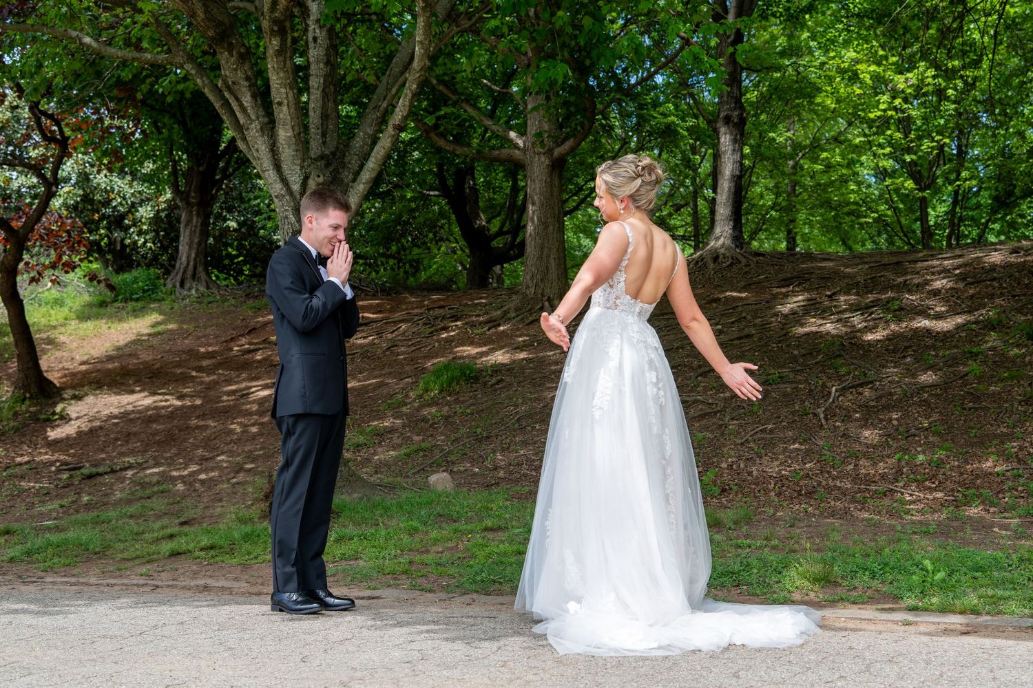 Bride in white gown and groom in black tuxedo share a first look moment on a tree-lined path.