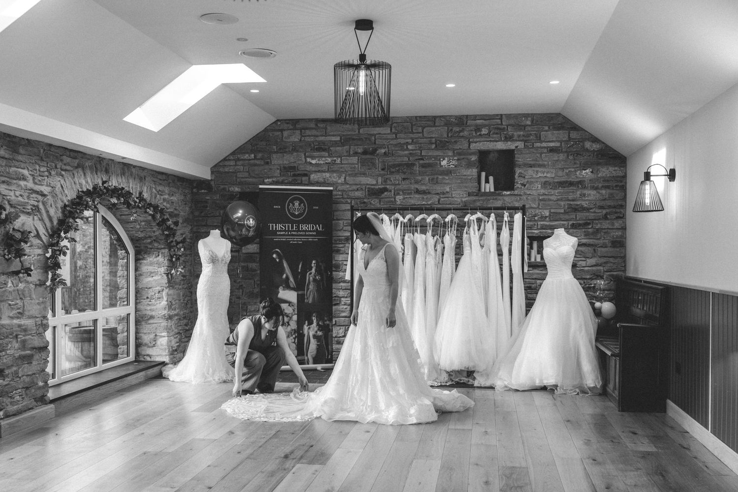 Black and white photo of a bridal boutique with wedding gowns on display and a consultant adjusting a bride's dress.