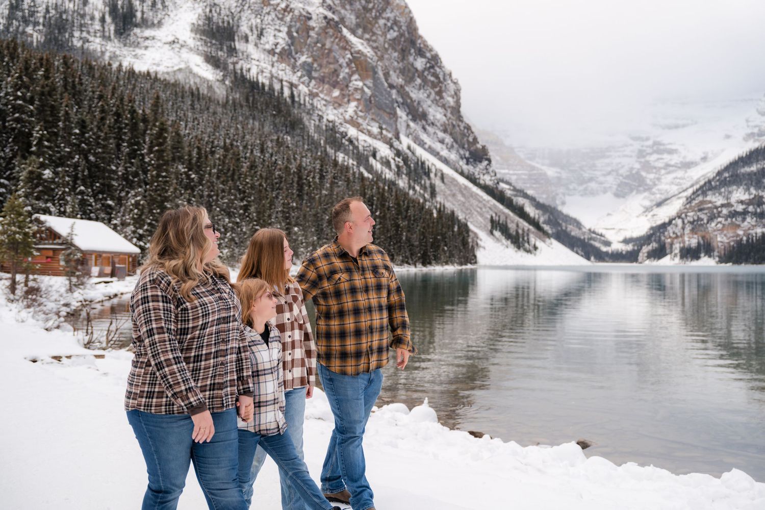 Family in flannel shirts poses by snowy Lake Louise with mountain backdrop in winter.