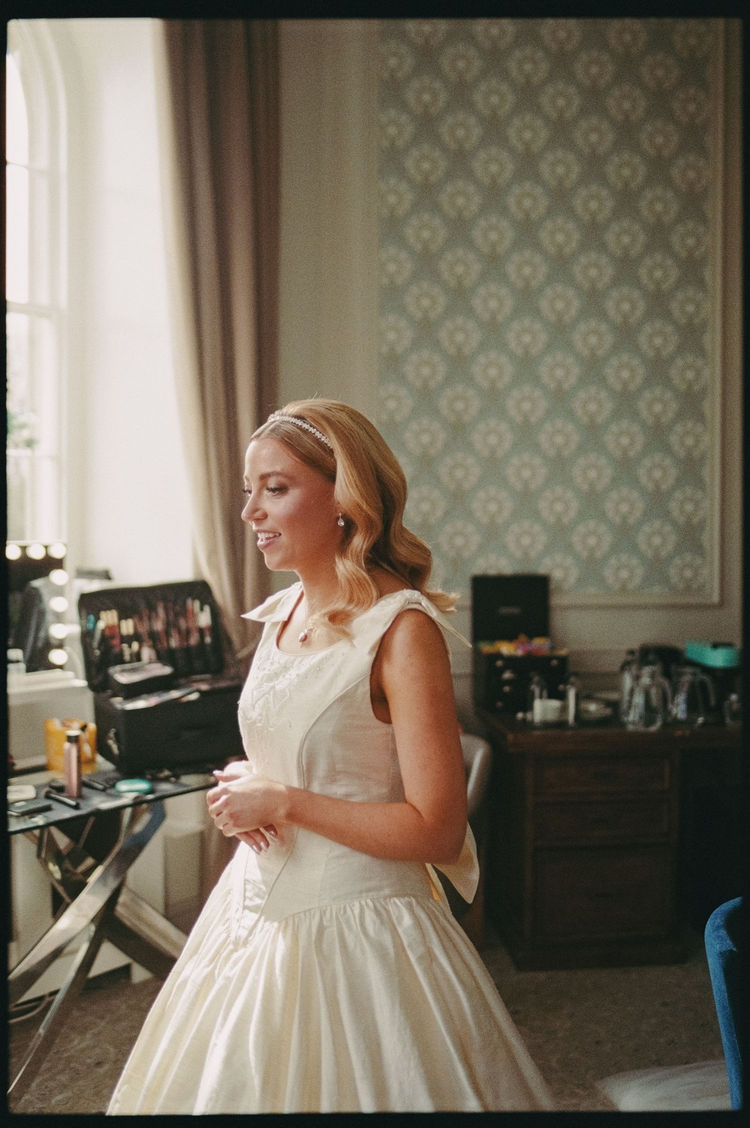 Blonde bride in vintage-style ivory wedding dress standing in elegant dressing room with patterned wallpaper.