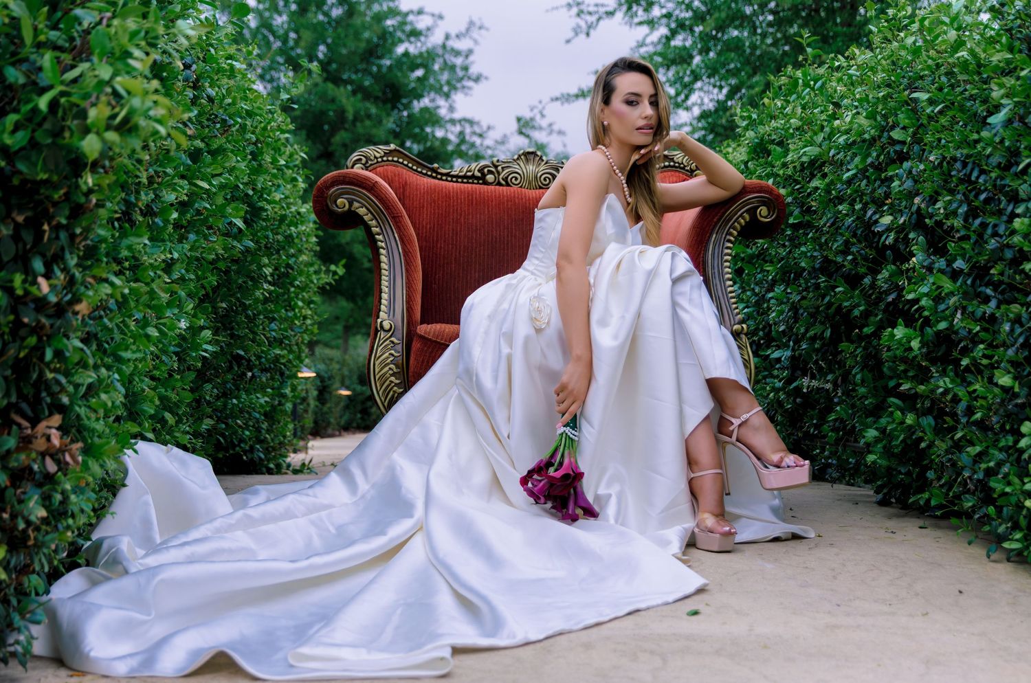 Bride in a white gown with train sits on a vintage red sofa surrounded by lush green hedges, holding a pink bouquet.