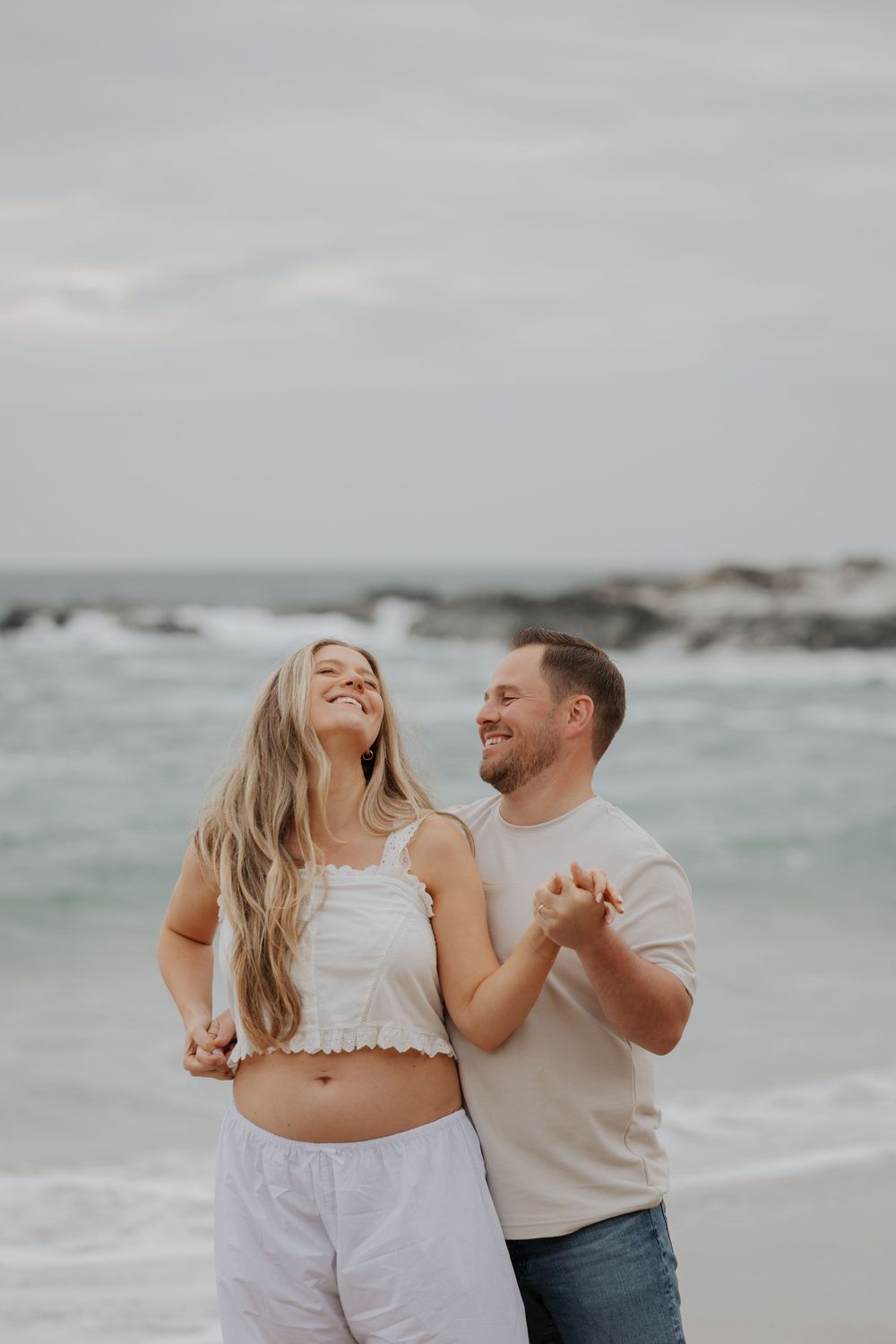 Joyful pregnant couple dancing together on a beach, woman laughing with long blonde hair flowing. Cornwall Maternity Photographer