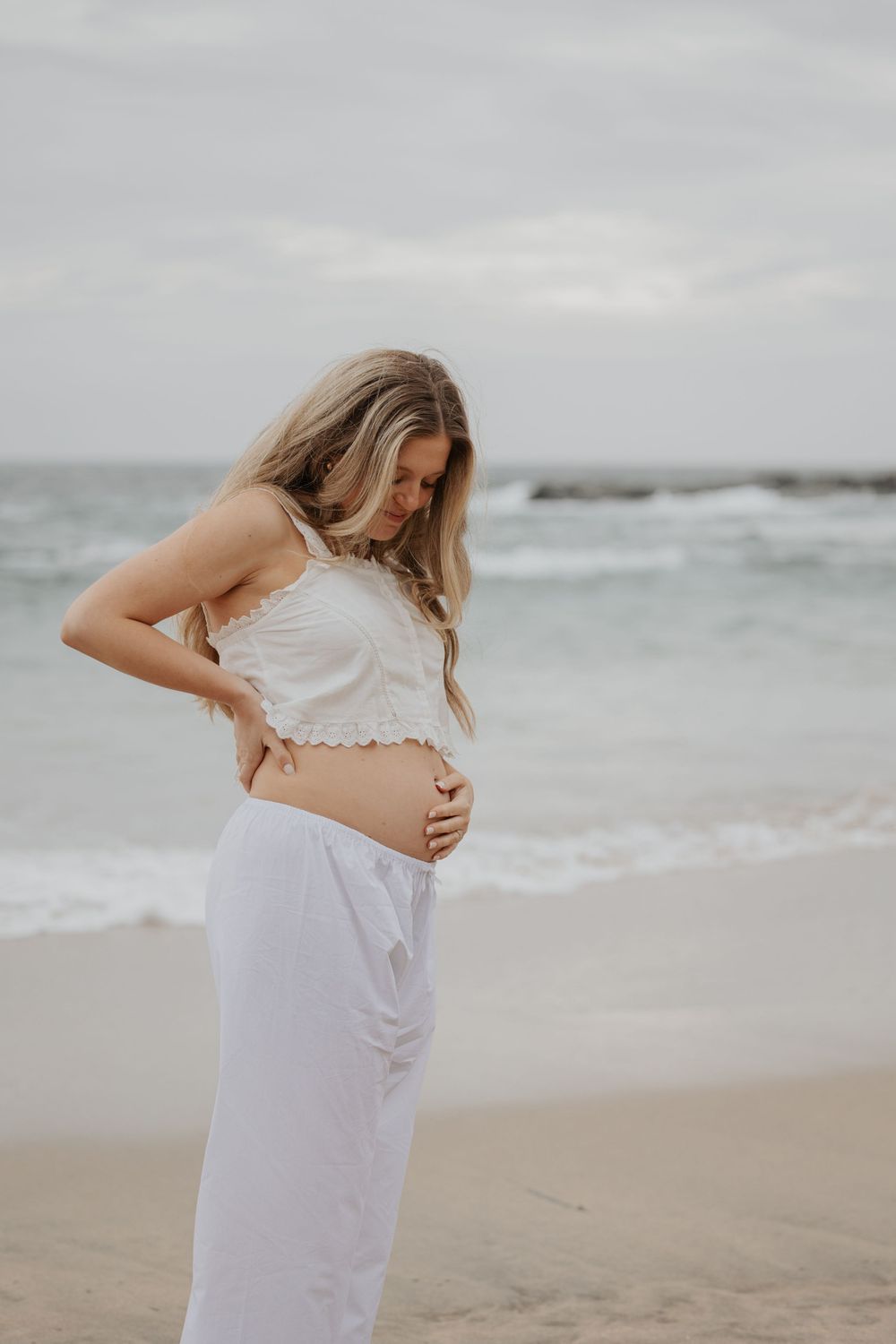 Pregnant blonde woman in white crop top and pants cradles her bare baby bump on a cloudy beach. Cornwall Maternity Photographer
