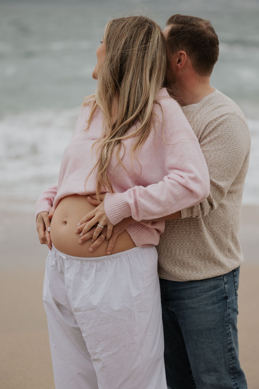 Couple embracing on beach, man kissing pregnant partners head while holding her bare baby bump tenderly. Cornwall Maternity Photographer