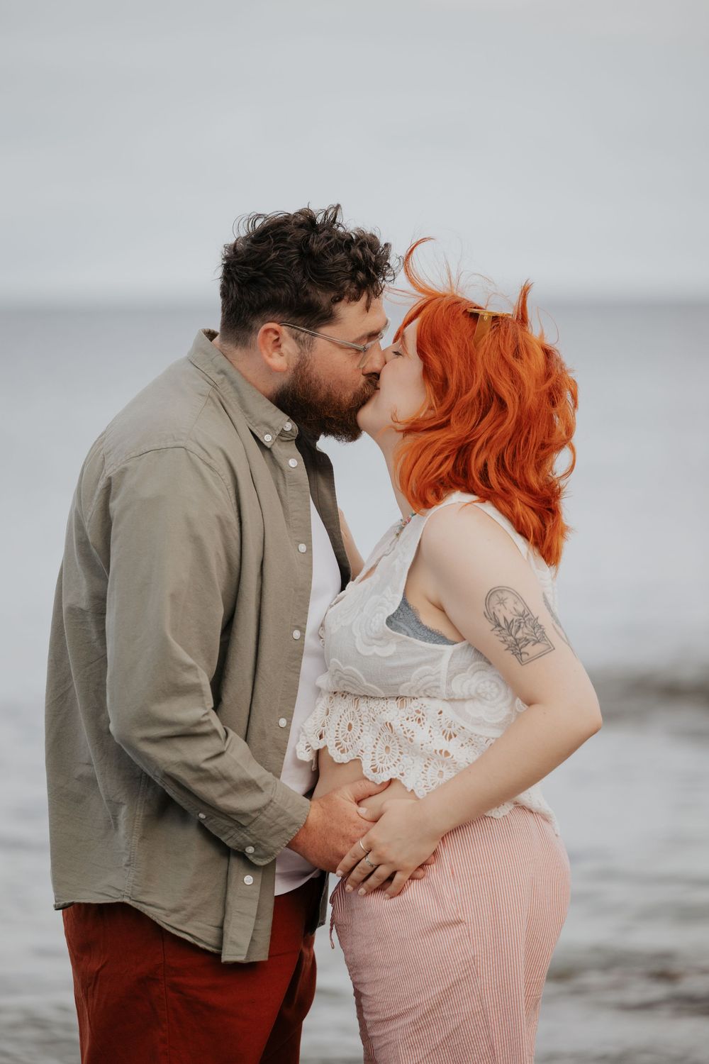 Couple sharing a kiss on beach, red-haired pregnant woman in white lace top holding partners hands. Cornwall Maternity Photographer