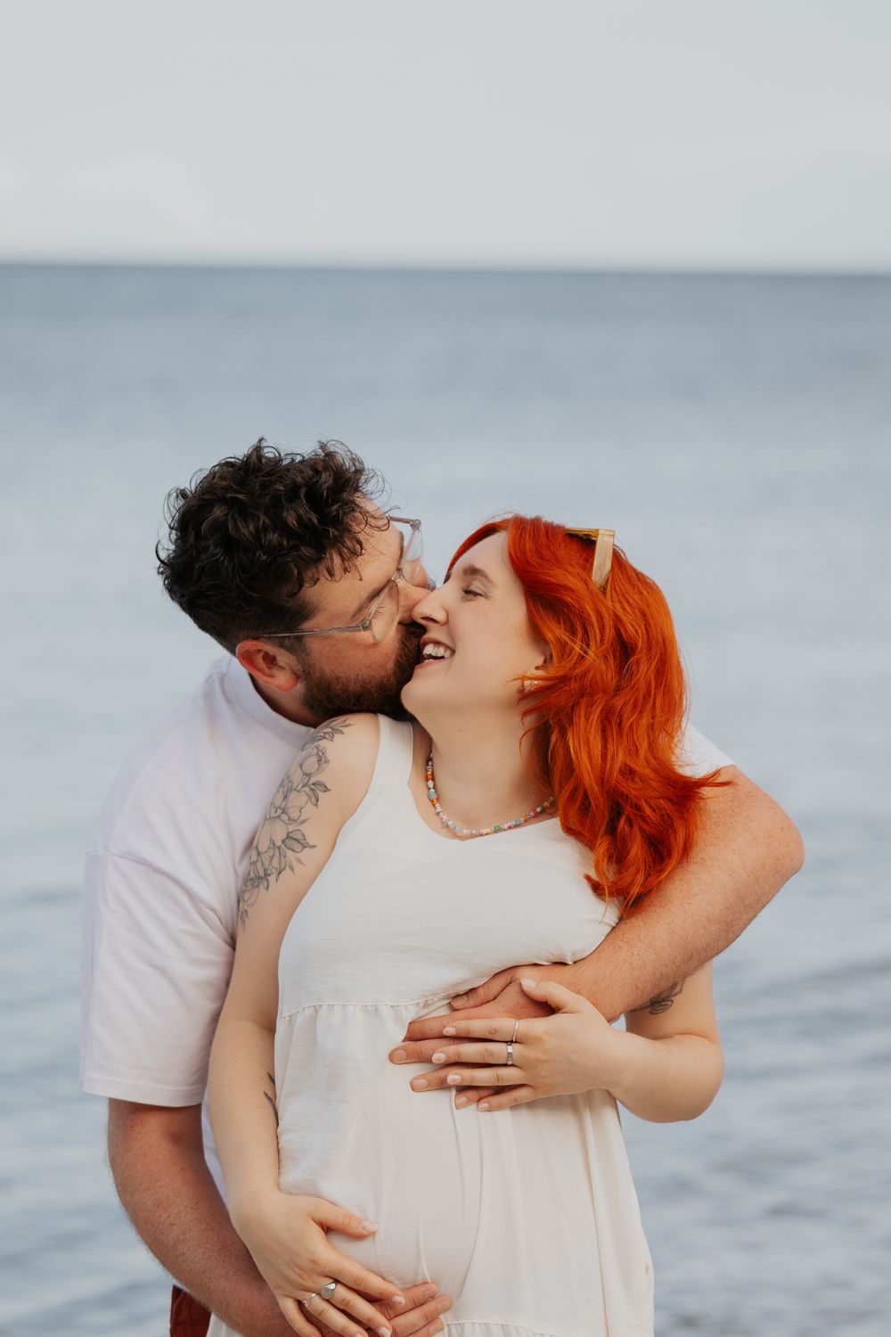 Bearded man nuzzling smiling red-haired pregnant woman in white dress on calm ocean beach backdrop. Cornwall Maternity Photographer