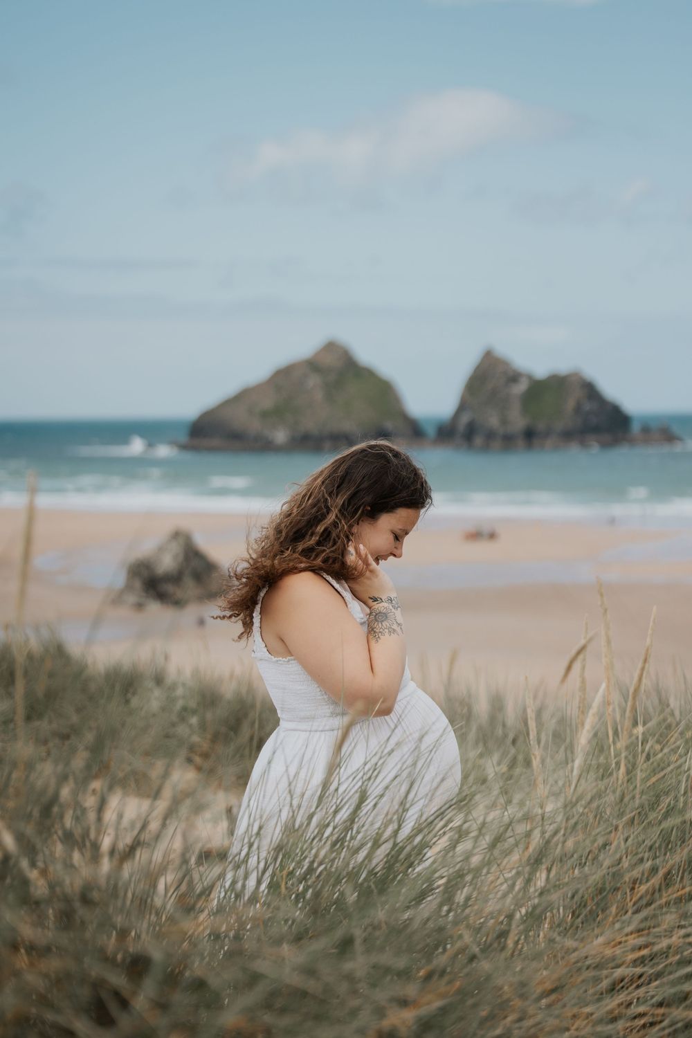 Pregnant woman in white dress standing in beach grass dunes with ocean and rocks in background. Cornwall Maternity Photographer