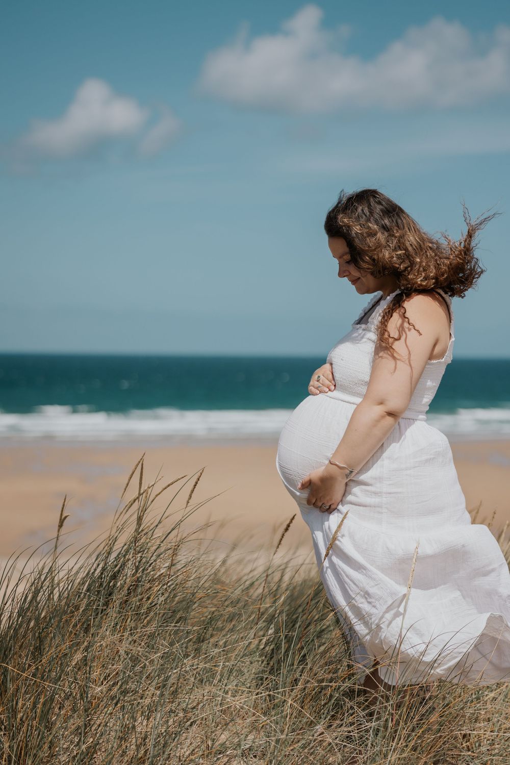 Side profile of pregnant woman in white dress cradling baby bump on sandy beach dunes. Cornwall Maternity Photographer
