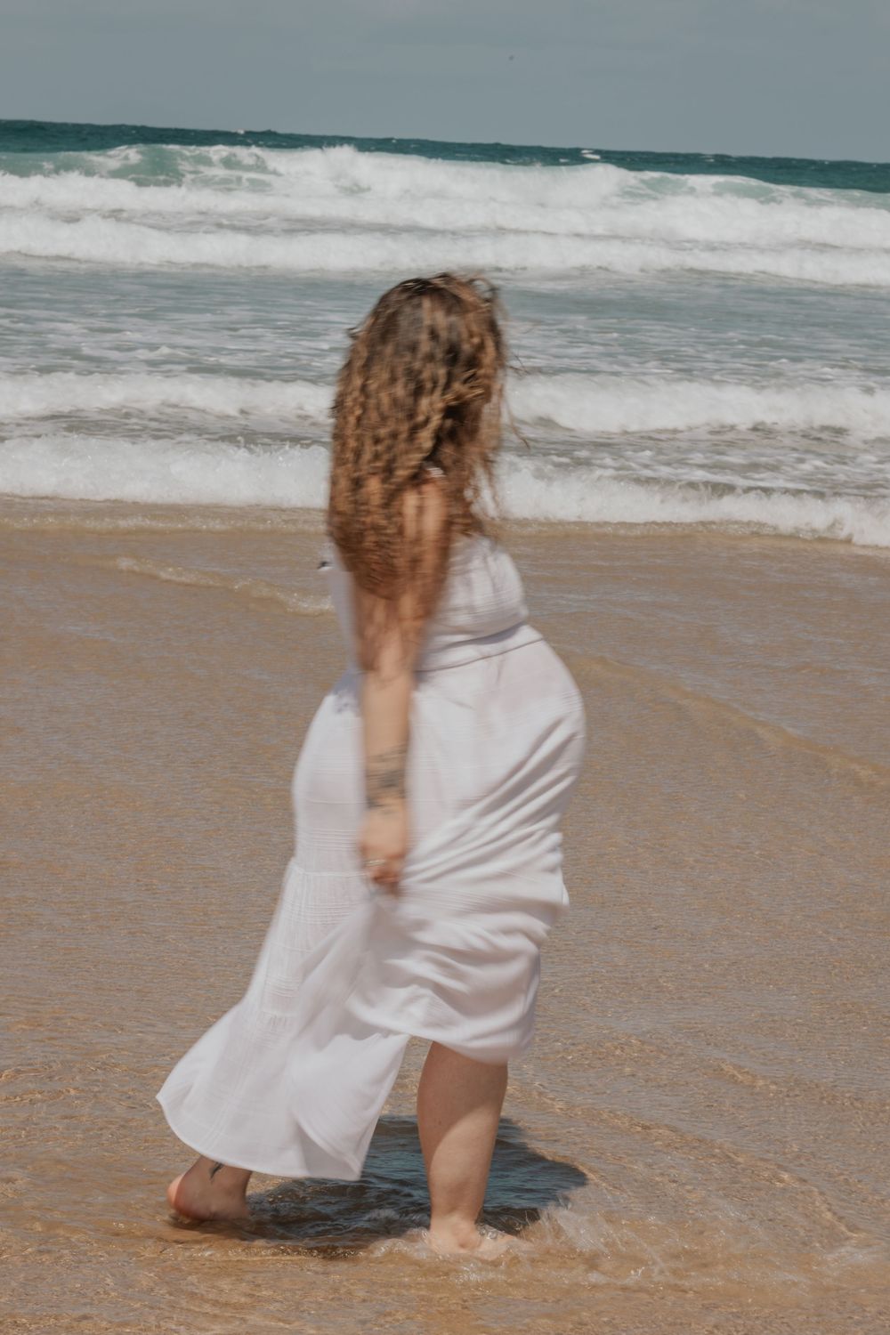 Pregnant woman in flowing white dress walking barefoot in ocean waves on sandy beach. Cornwall Maternity Photographer
