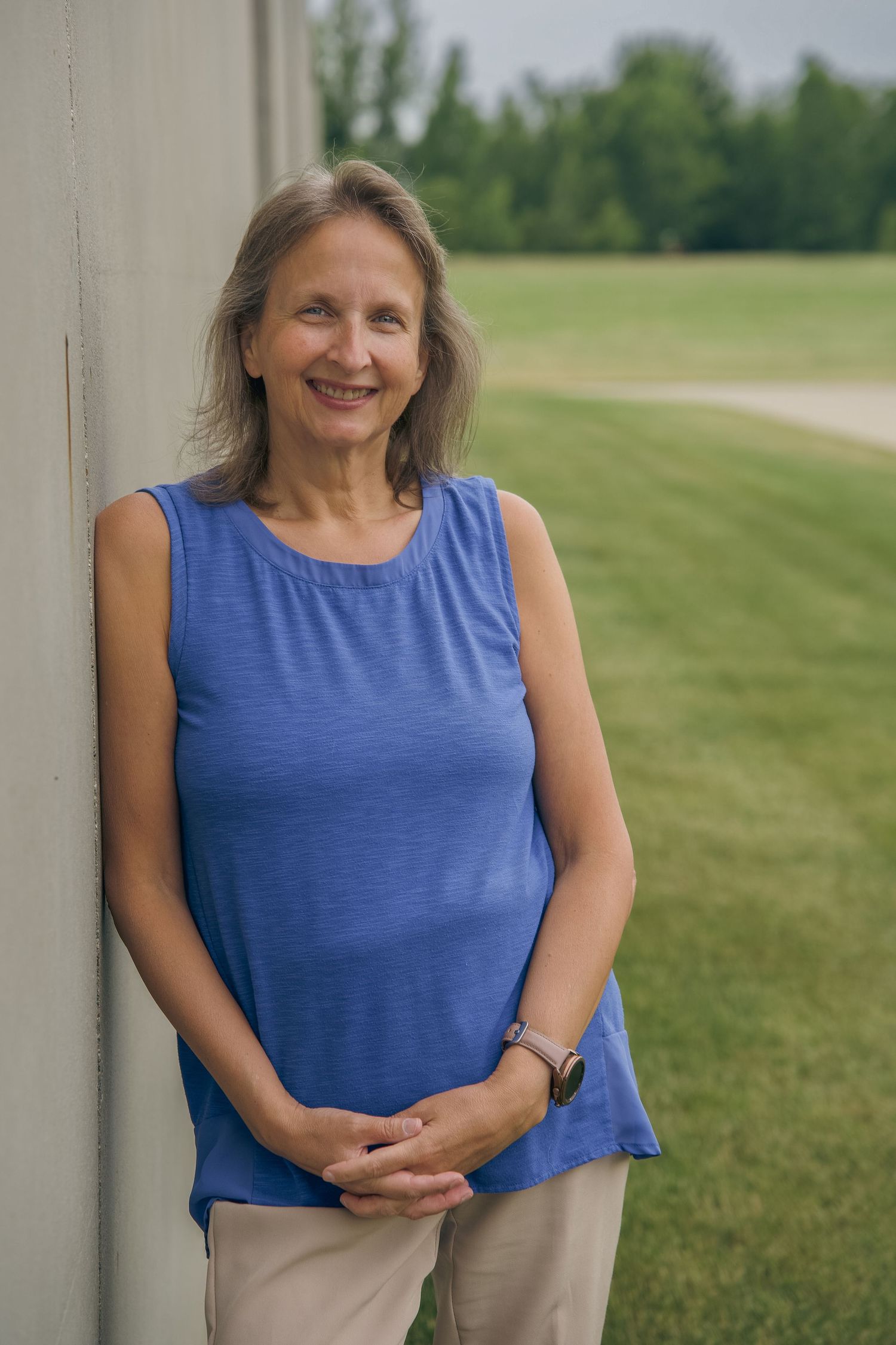 Smiling middle-aged woman with gray hair leaning against a white wall outdoors, wearing a blue sleeveless top.