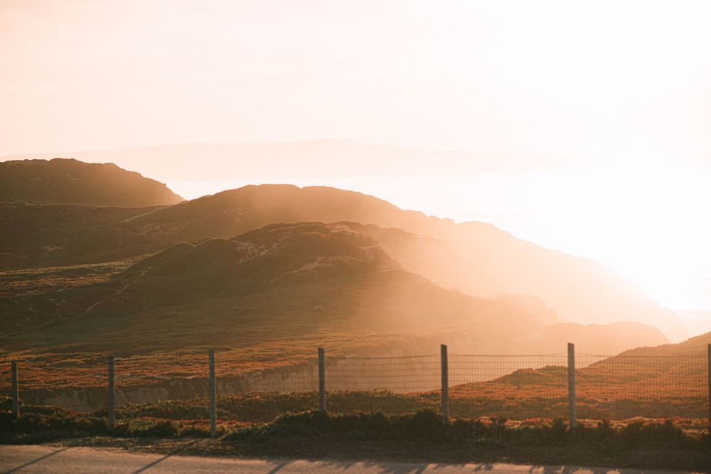 Golden sunlight illuminates rolling hills and mountains behind a wire fence in a serene rural landscape at sunset.