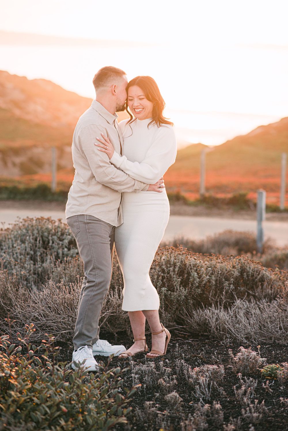 Couple embracing at golden hour sunset in coastal landscape, wearing neutral tones, smiling warmly.