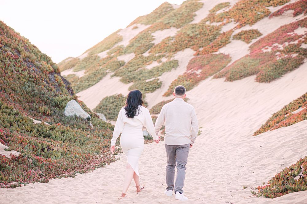 Couple holding hands walking up sandy dunes with colorful coastal vegetation during an engagement photo session.