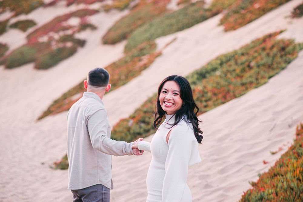 Couple holding hands on sandy beach dunes, woman in white dress smiling over shoulder, colorful coastal plants in background.