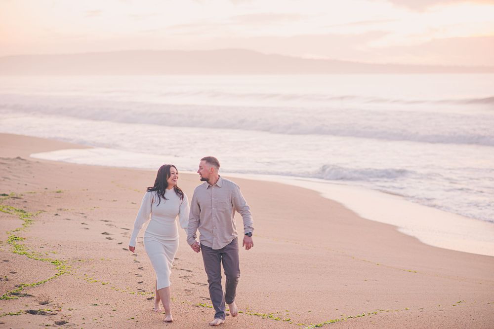 Couple walking hand-in-hand on a sandy beach at sunset, smiling at each other with gentle waves in the background.