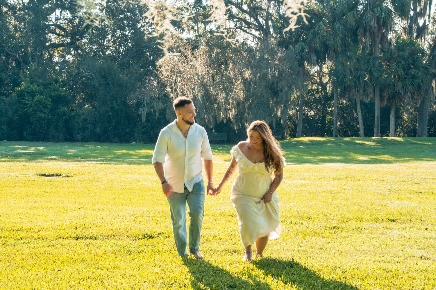 Pregnant couple holding hands walking on lush green grass surrounded by moss-draped trees in soft sunlight.