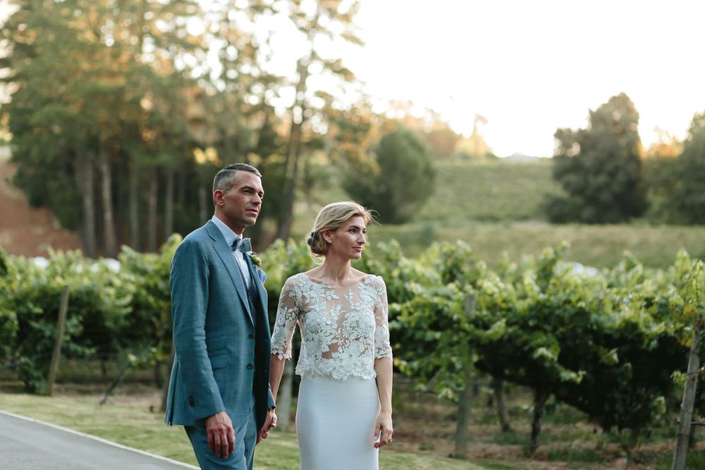 Eine Braut in einem Brautkleid mit Blumenspitzen und ein Bräutigam in einem blauen Anzug und Weinreben im Hintergrund