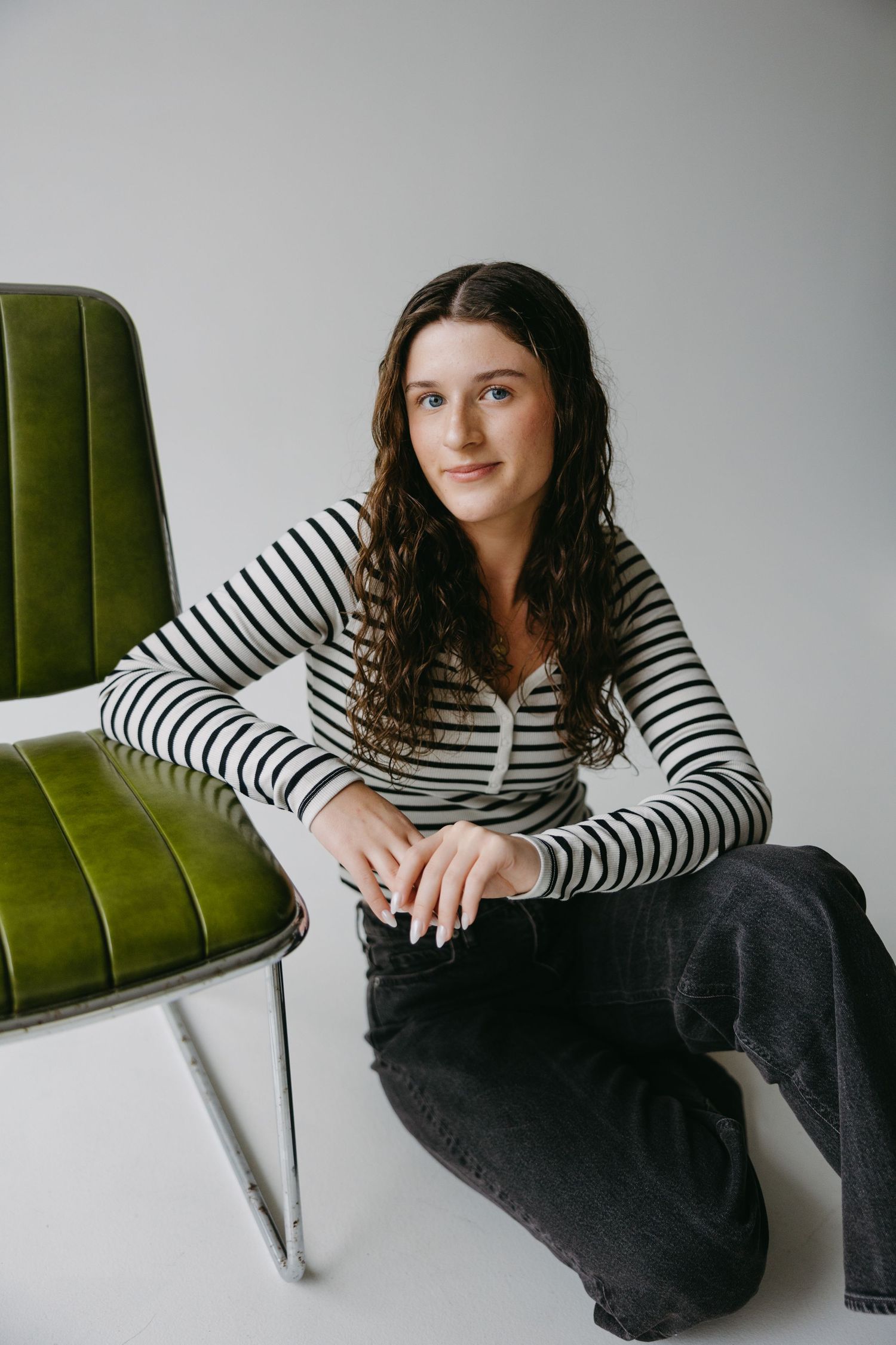 Person in striped shirt sitting beside green velvet chair against white wall.