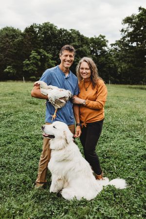 couple holding chicken with big white dog on their farm