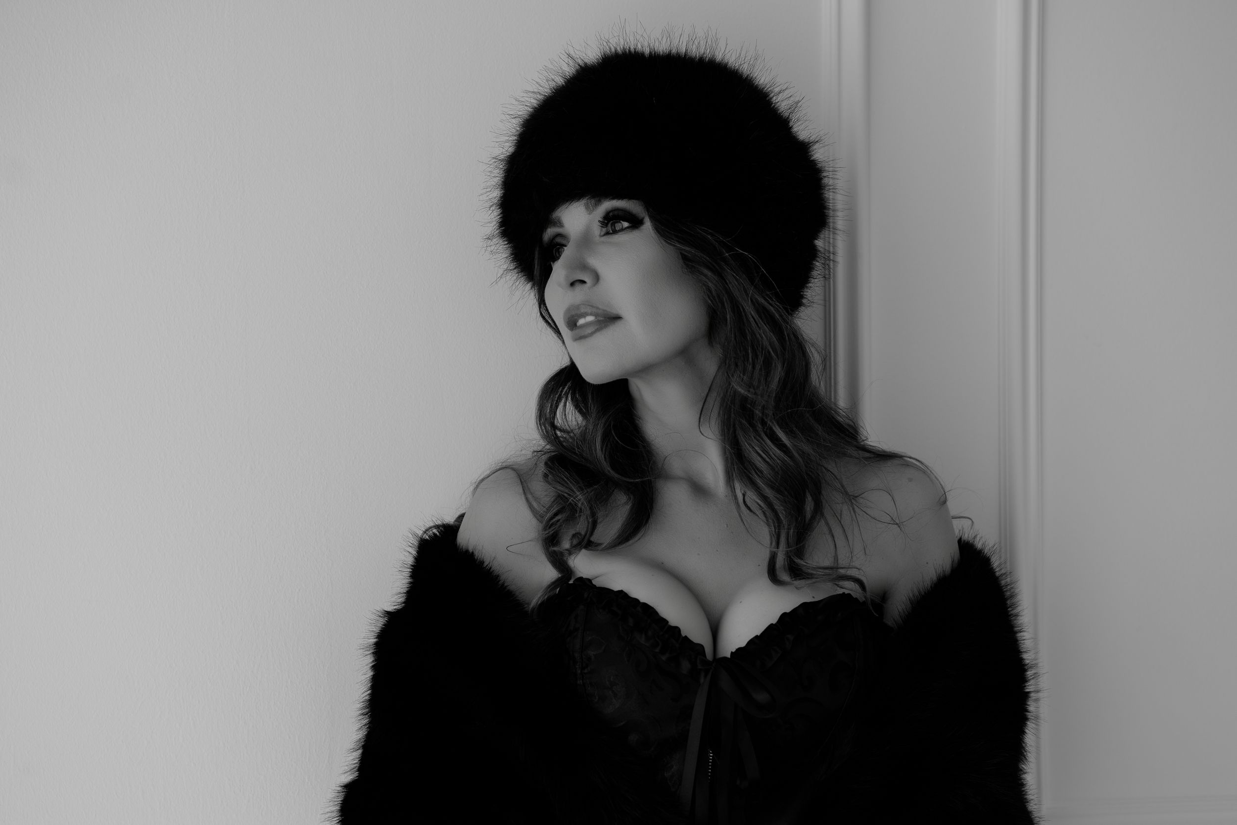 Black and white photo of a woman in a fur hat and coat posing elegantly against a light wall.