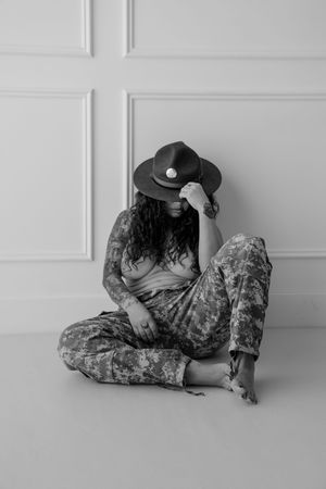 A person in military fatigues sits cross-legged on a white floor while wearing a dark cap against a bright wall.