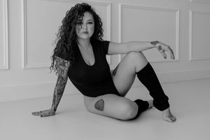 Black and white portrait of a person in athletic wear posing on floor with arm tattoo and legwarmers.