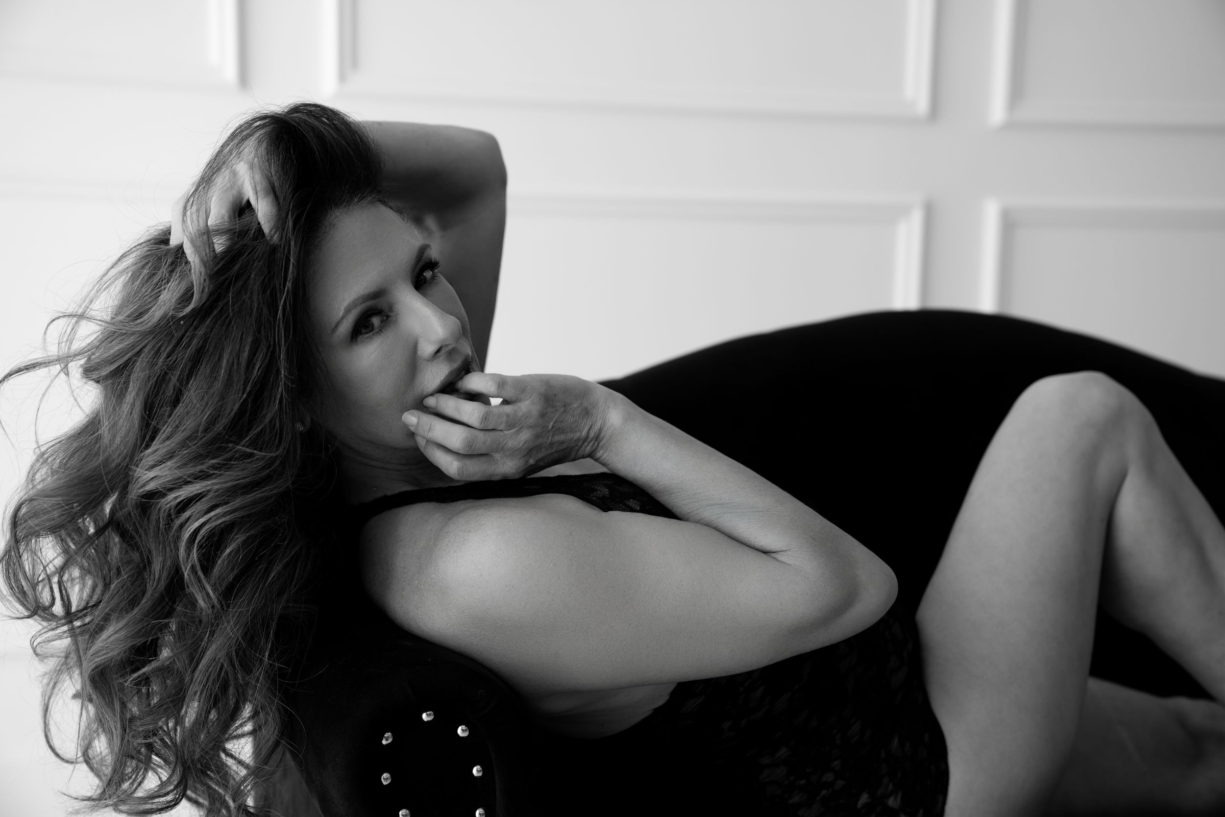 Black and white portrait of a woman with wavy hair reclining on dark furniture in an artistic pose.