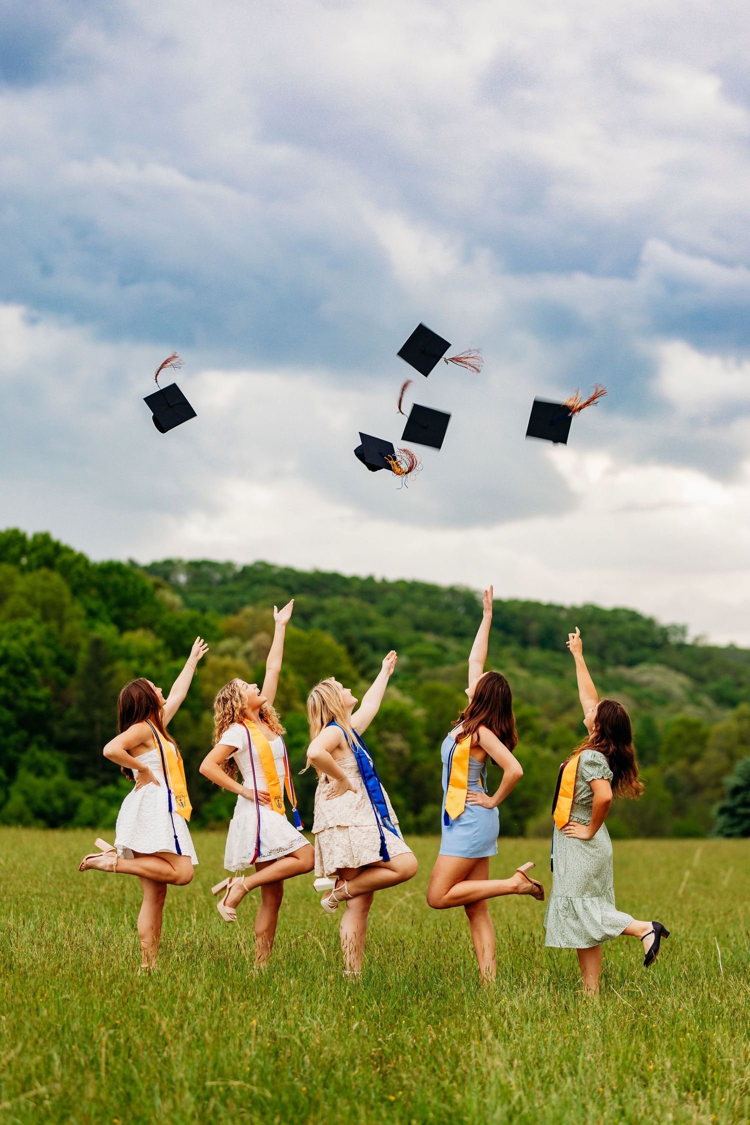 Five female graduates tossing caps in green field under cloudy sky celebrating graduation.