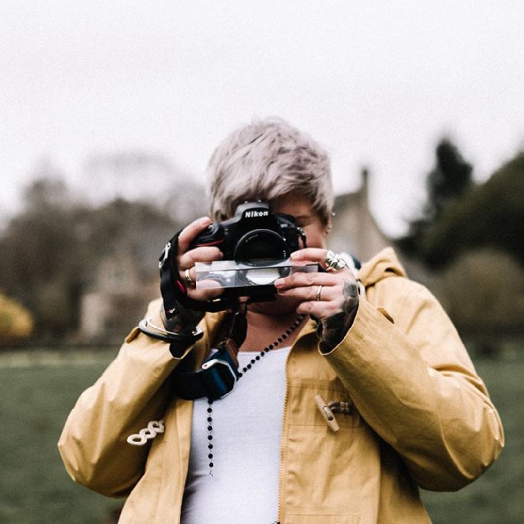Person in yellow jacket taking photo with professional camera against blurred outdoor background.