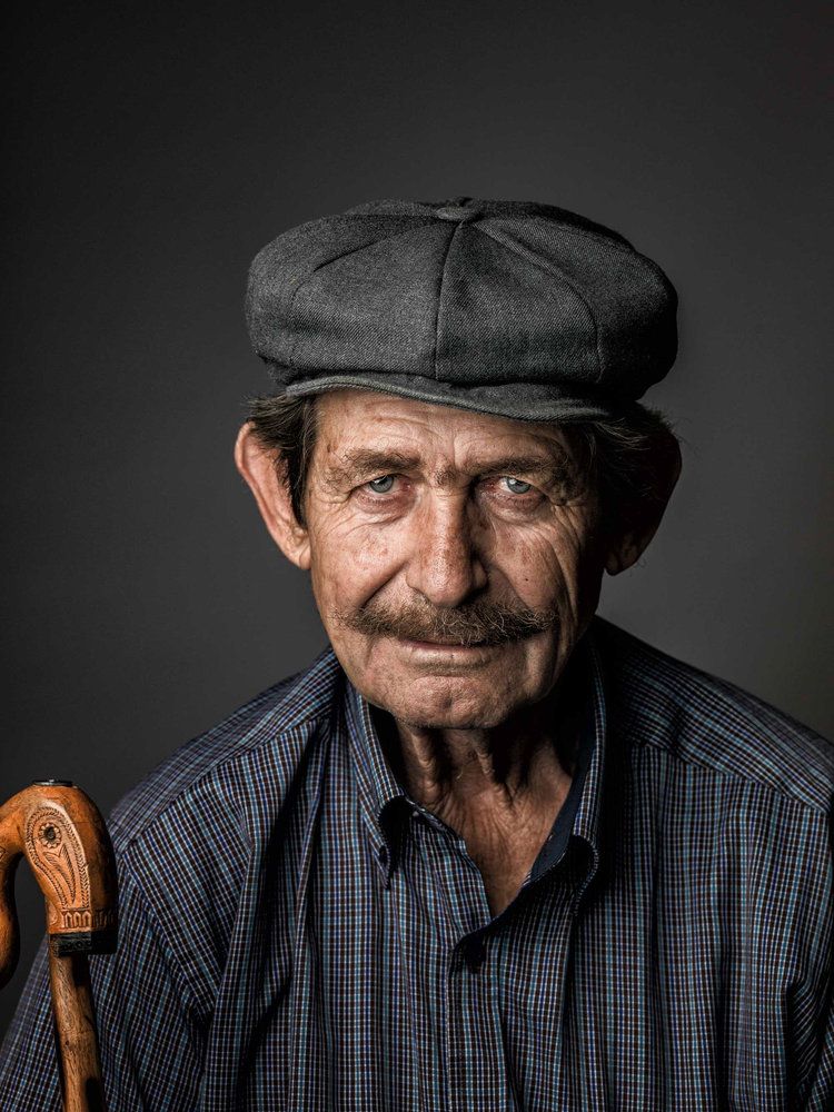 Studio portrait of a person in a dark cap and plaid shirt holding a wooden cane.