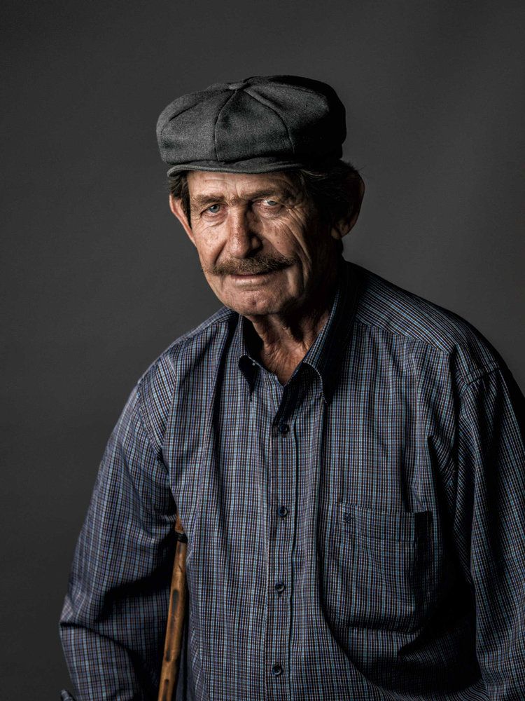Portrait of an elderly figure wearing a flat cap and plaid shirt against dark background.