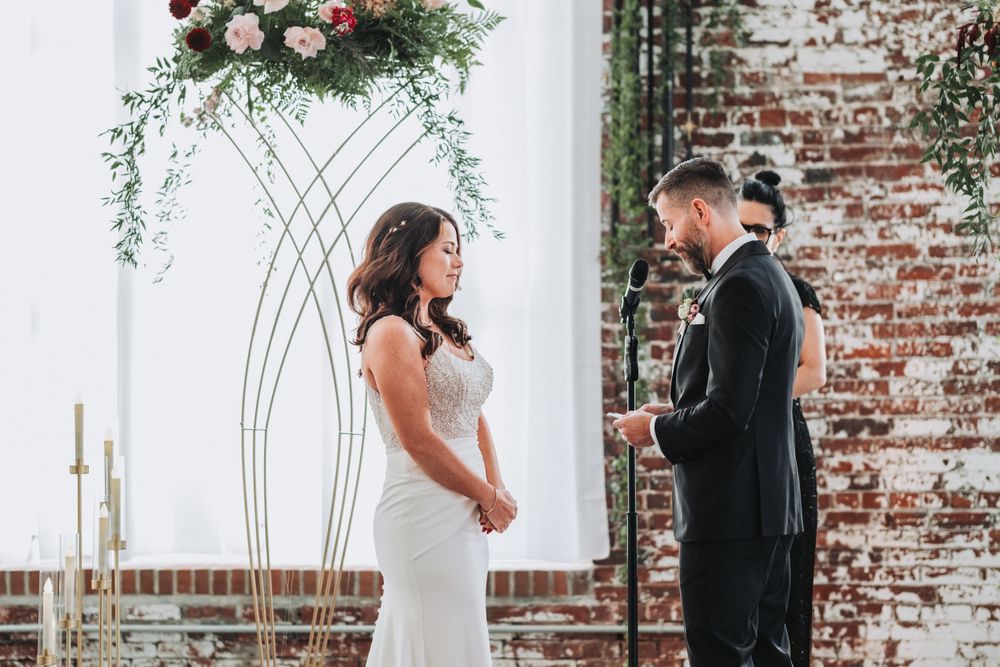 Couple exchanges vows during wedding ceremony under floral arch against exposed brick wall.