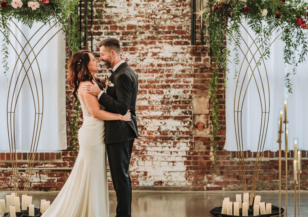 Couple embraces in front of rustic exposed brick wall decorated with geometric panels and greenery.