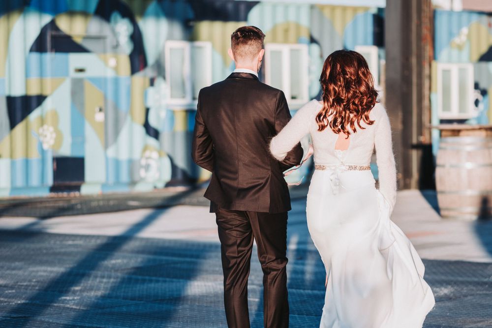 A couple in wedding attire walking together down a sunlit corridor with bright light streaming through windows.