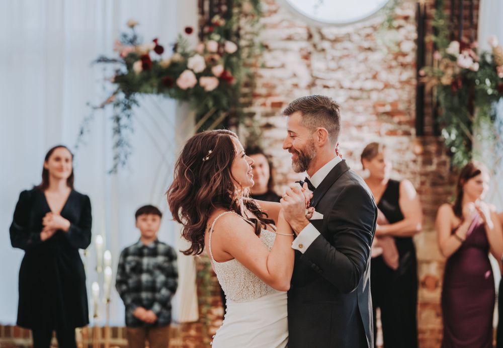 A romantic first dance at a rustic brick-walled venue decorated with floral arrangements and white drapery.