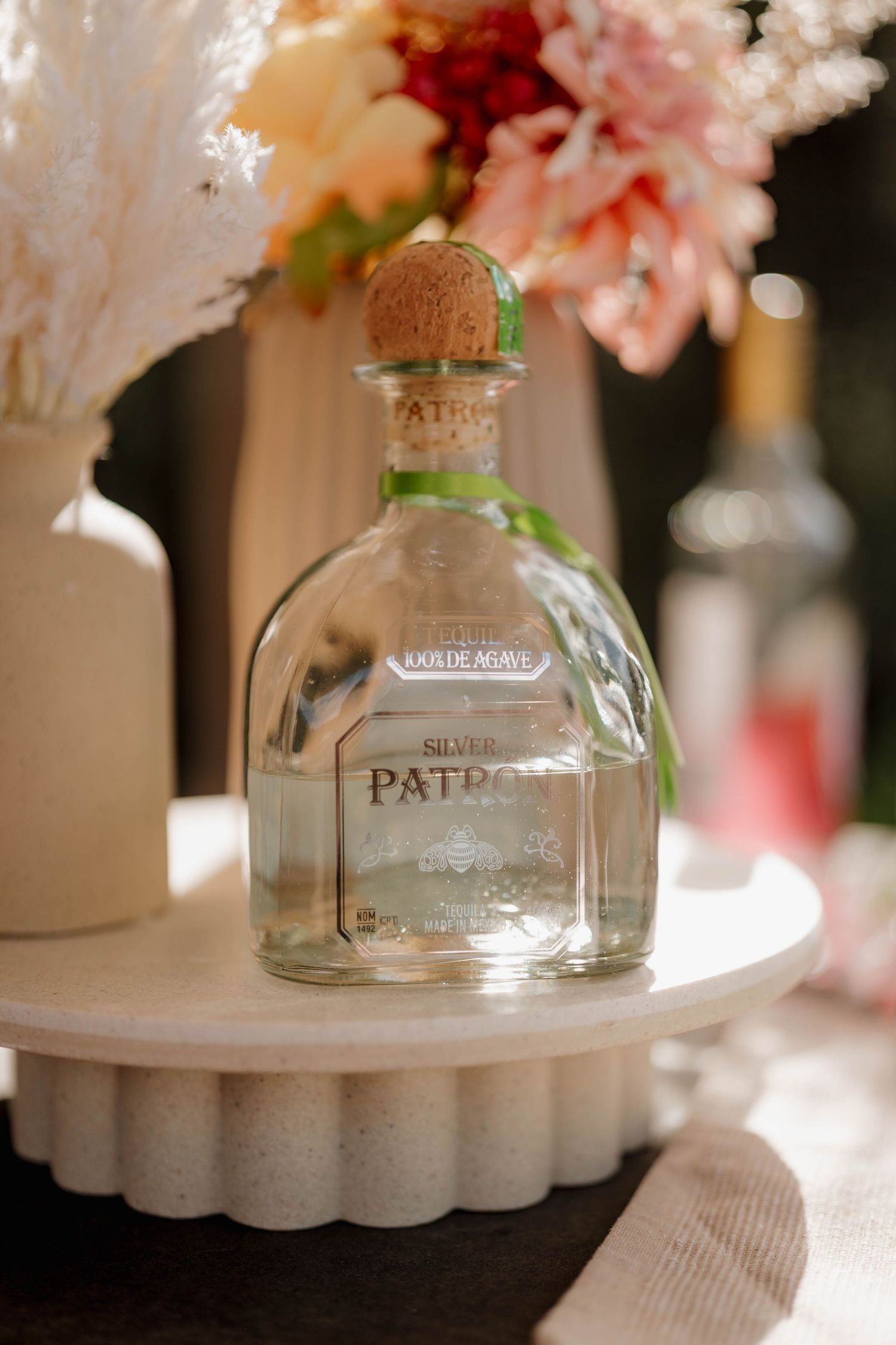 Patron Silver tequila bottle with cork stopper on white cake stand, surrounded by pink and white floral arrangements.