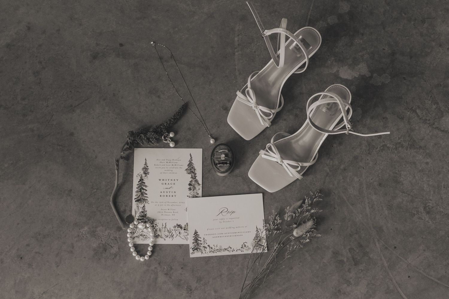 Black and white flat lay of bridal details including heels, wedding invitations, jewelry, and accessories on textured floor.