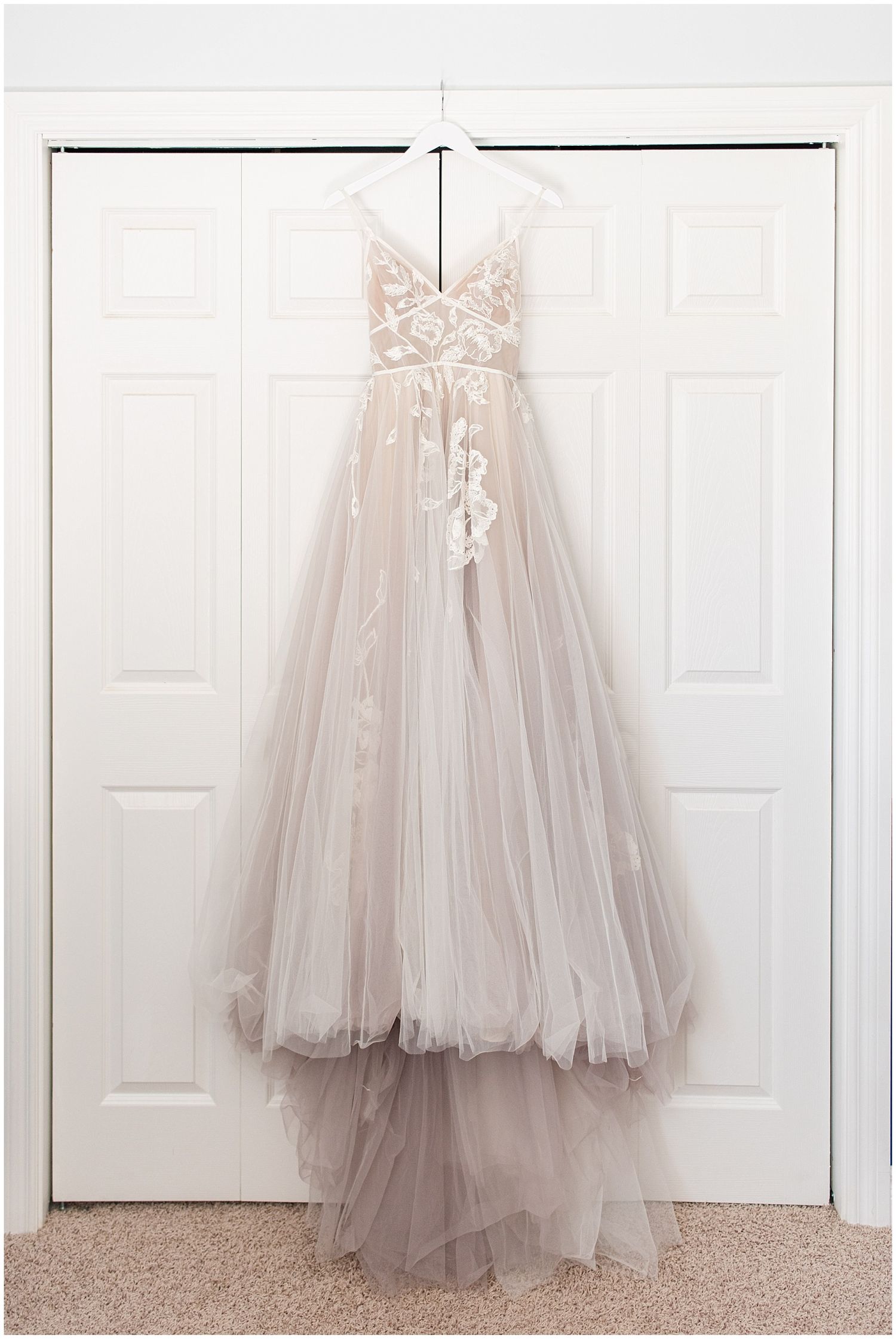 Elegant blush tulle wedding gown with floral lace bodice hanging on white closet doors.