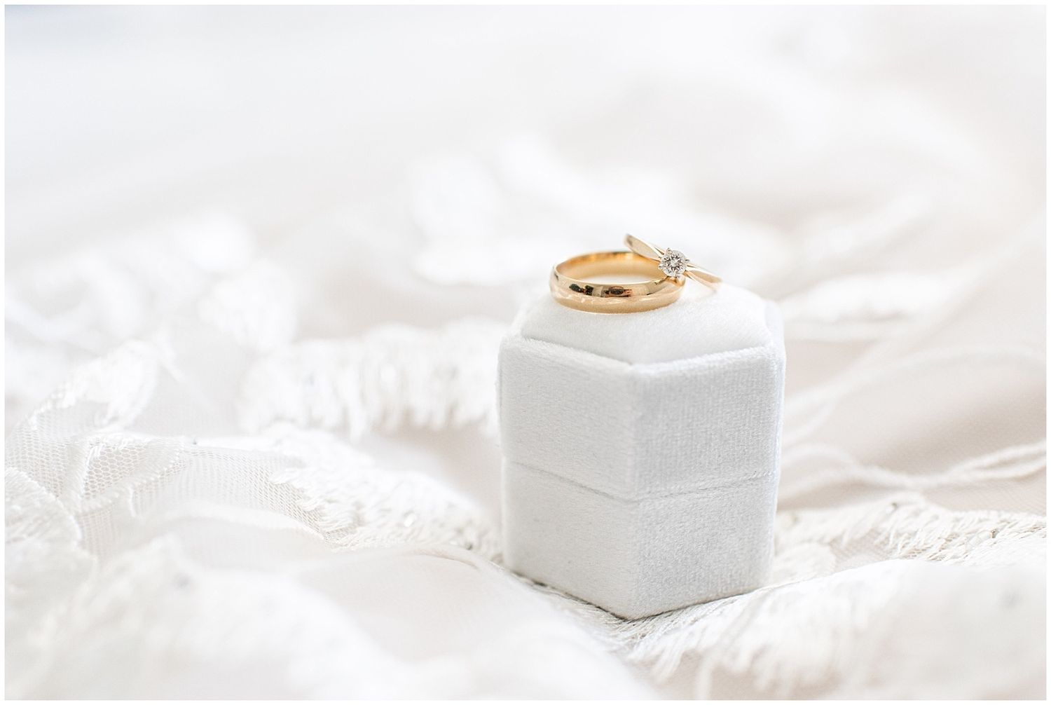 Gold wedding bands and diamond engagement ring displayed on a white velvet ring box atop soft white fabric.