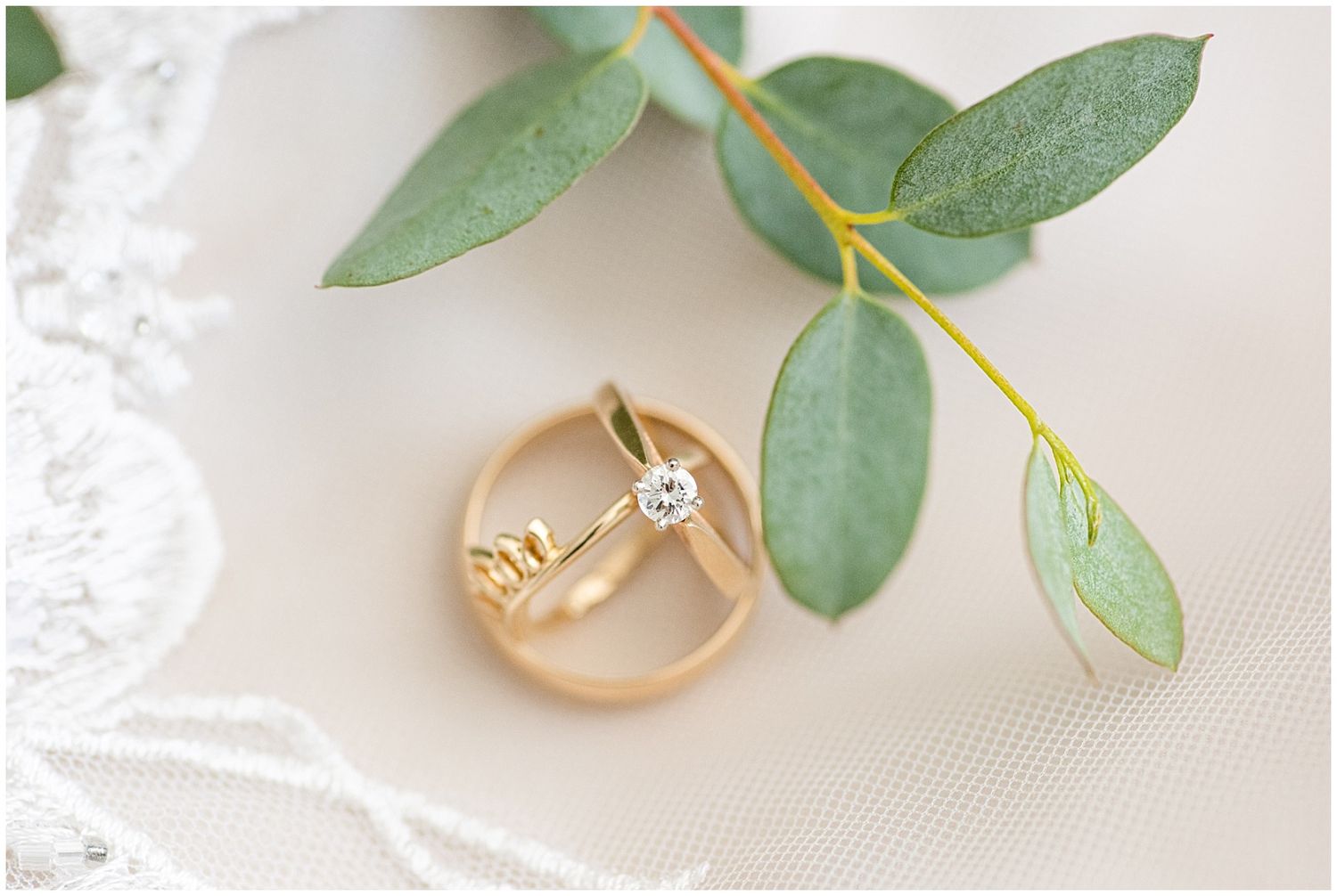 Gold wedding bands and diamond engagement ring resting on white lace with eucalyptus greenery backdrop.