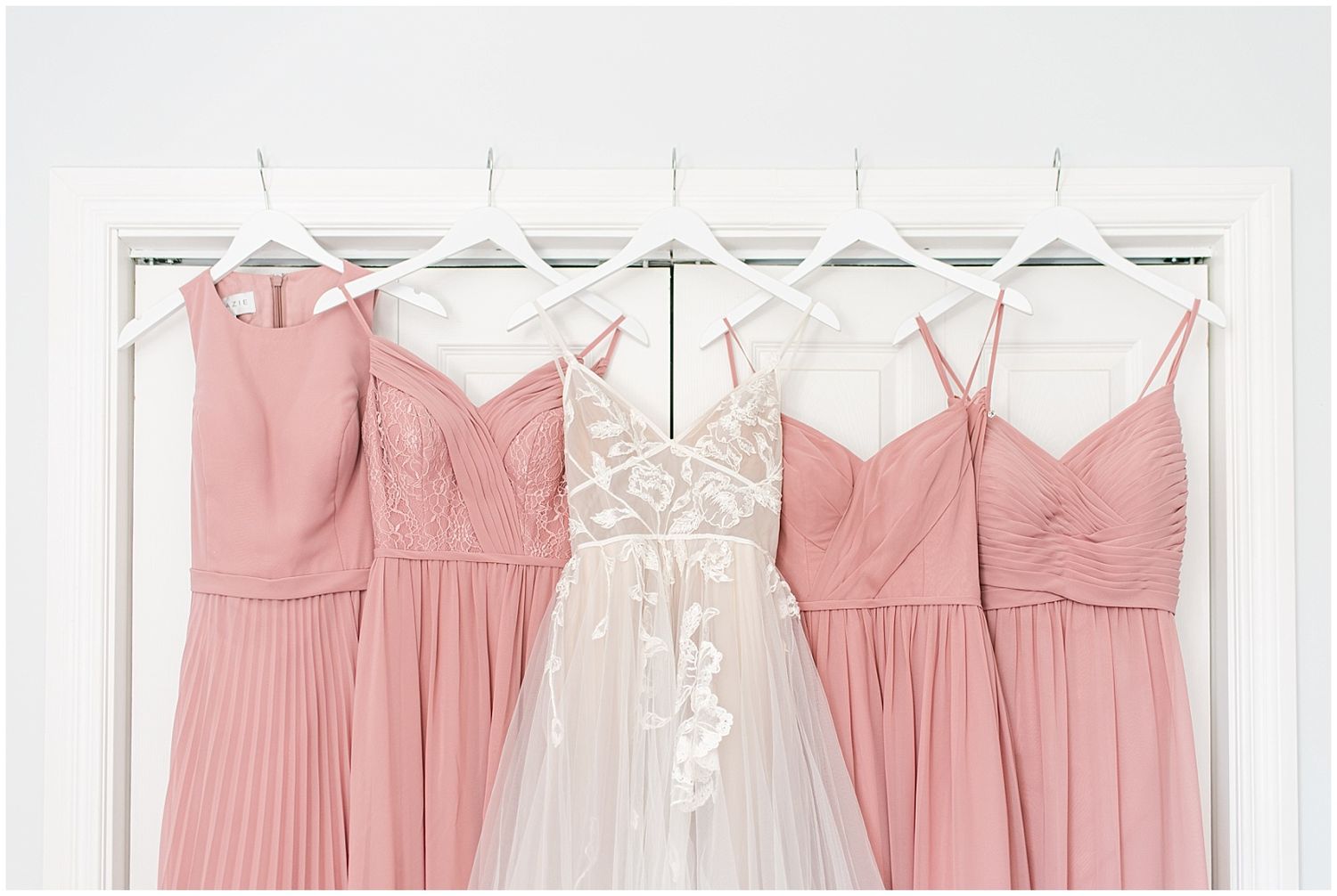 White lace wedding gown flanked by four blush pink bridesmaid dresses hanging on white hangers against a white door.