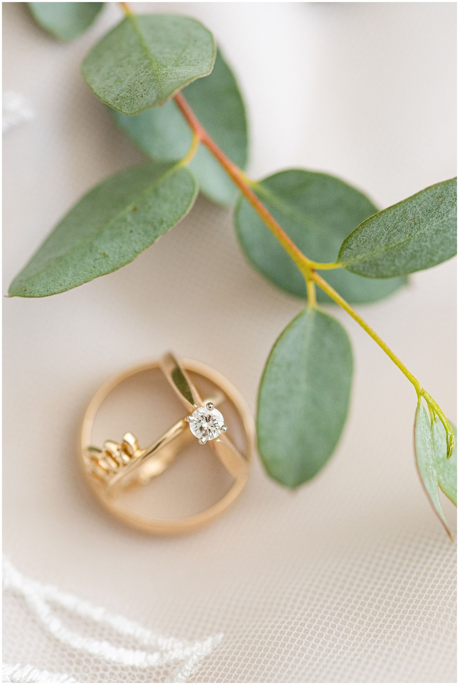 Gold engagement ring and wedding band with diamond solitaire resting against eucalyptus leaves on white fabric.
